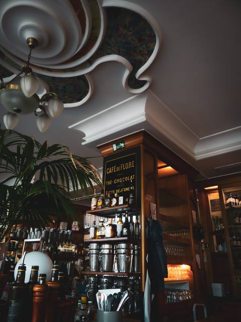 Die Bar unter der geschwungenen Stuckdecke: Kronleuchter, Palme, Flaschenregal und das Schild „Café de Flore“