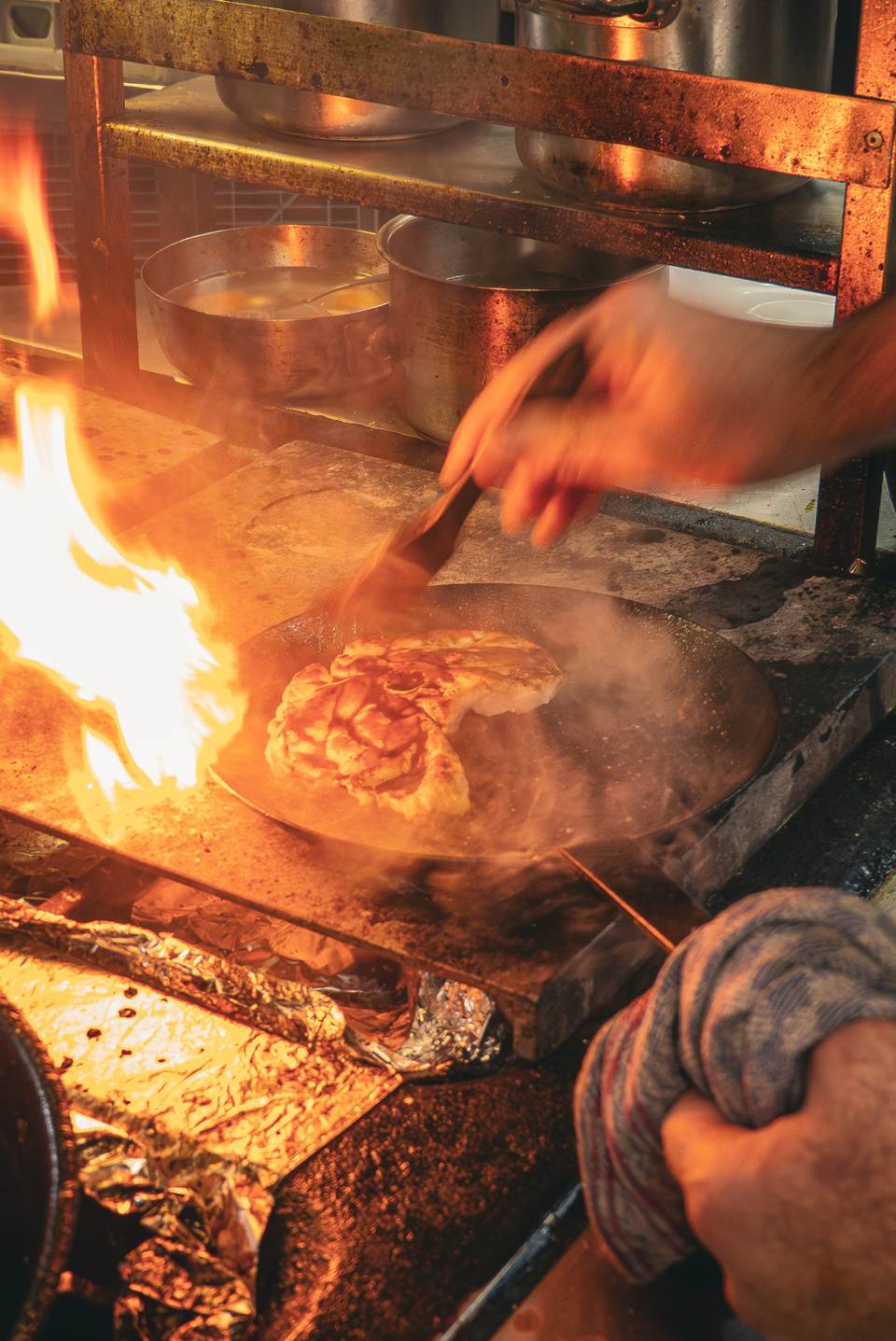 Offenes Feuer in der Küche: eine Hand wendet Fleisch in der Pfanne, Flammen schlagen hoch
