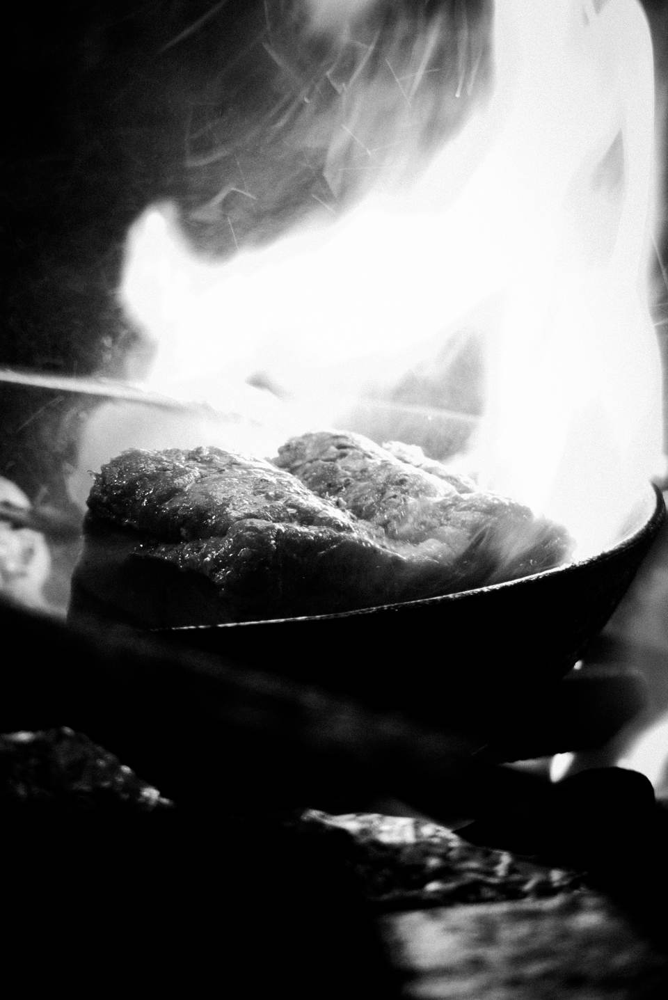 Schwarzweiß, ganz nah: Fleisch in der Pfanne über offenem Feuer, Flammen und Rauch dahinter
